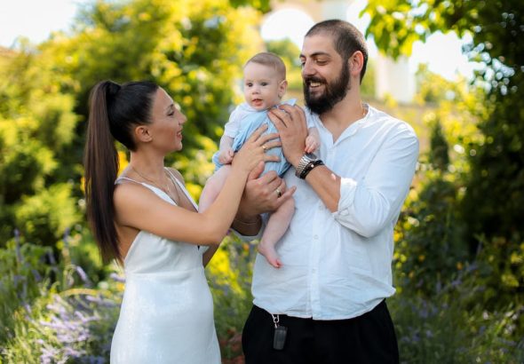 Mother and father joyfully hold their baby in a vibrant outdoor garden setting.