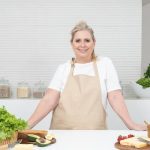 A mature woman in an apron stands at a kitchen counter with fresh produce and cheese, ready for cooking.