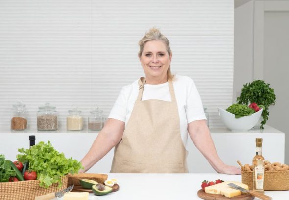 A mature woman in an apron stands at a kitchen counter with fresh produce and cheese, ready for cooking.
